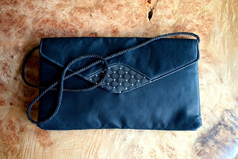 Puede incluir: Un clutch de sat&eacute;n negro con una correa de cuerda a juego. La solapa presenta un panel con dise&ntilde;o de diamantes con peque&ntilde;os adornos dorados. El bolso rectangular es adecuado para ocasiones formales.