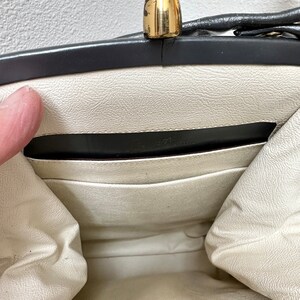 Puede incluir: Vista abierta de un bolso vintage con un cierre dorado. El interior es blanco roto con un ribete negro y un peque&ntilde;o bolsillo. El interior del bolso es visible, mostrando el forro y los detalles de construcci&oacute;n. El cierre es una caracter&iacute;stica destacada.