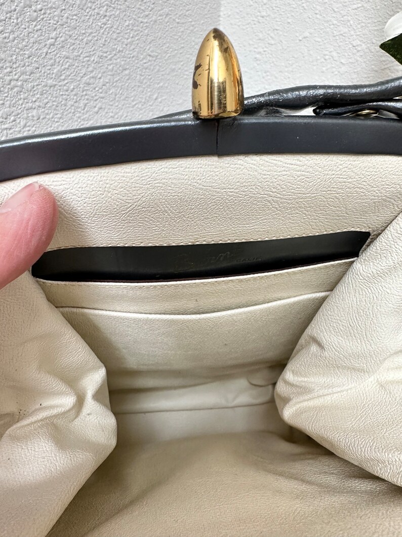 Puede incluir: Vista abierta de un bolso vintage con un cierre dorado. El interior es blanco roto con un ribete negro y un peque&ntilde;o bolsillo. El interior del bolso es visible, mostrando el forro y los detalles de construcci&oacute;n. El cierre es una caracter&iacute;stica destacada.