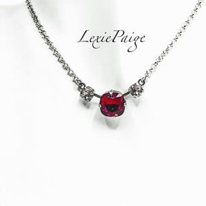May include: A silver-toned necklace featuring a large, square, ruby-red crystal pendant, flanked by two smaller, clear crystal accents. The necklace has a delicate chain and the text "Lexie Paige" is visible.