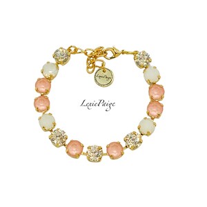 Puede incluir: Un brazalete dorado con una cadena y cristales redondos rosas y blancos alternados. El brazalete tiene una pequeña etiqueta dorada con el texto "Lexie Paige" en ella.