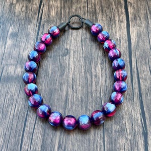Op de afbeelding: Een ketting met grote, ronde kralen in blauwe, paarse en roze tinten. De kralen zijn geregen met een zwarte metalen sluiting en connectoren. De ketting wordt gepresenteerd op een houten oppervlak.
