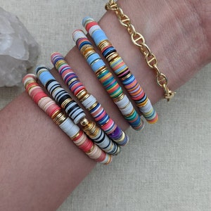 May include: A stack of colorful bracelets with gold accents. The bracelets are made of stacked, round, colorful beads with gold accents. The bracelets are worn on a person's wrist.