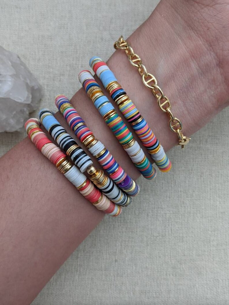 May include: A stack of colorful bracelets with gold accents. The bracelets are made of stacked, round, colorful beads with gold accents. The bracelets are worn on a person's wrist.