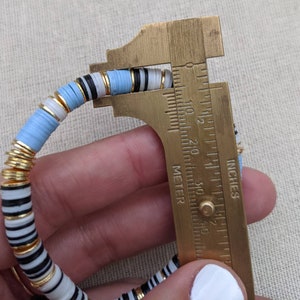 May include: A multi-colored bracelet with blue, white, black, and gold beads is measured with a caliper. The caliper reads 2 1/8 inches.