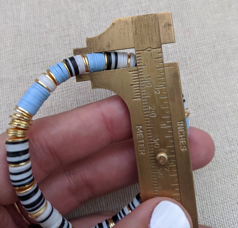 May include: A multi-colored bracelet with blue, white, black, and gold beads is measured with a caliper. The caliper reads 2 1/8 inches.
