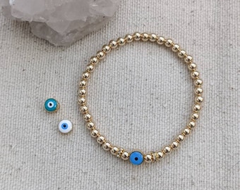 May include: Gold beaded bracelet with a blue evil eye charm. Two additional evil eye charms are next to the bracelet. The bracelet is made of small, round, gold-colored beads.