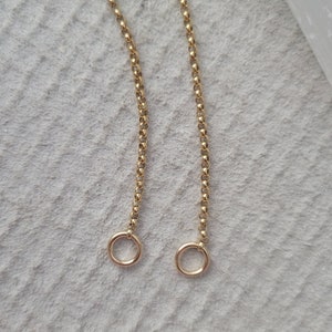 May include: Close-up of two gold-colored necklaces with delicate chain links and small circular pendants. The necklaces are displayed against a light gray, textured background, creating a simple and elegant aesthetic.