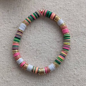 May include: A colorful beaded bracelet with pink, green, white, and yellow beads. The beads are separated by gold spacers. The bracelet is on a white fabric background.