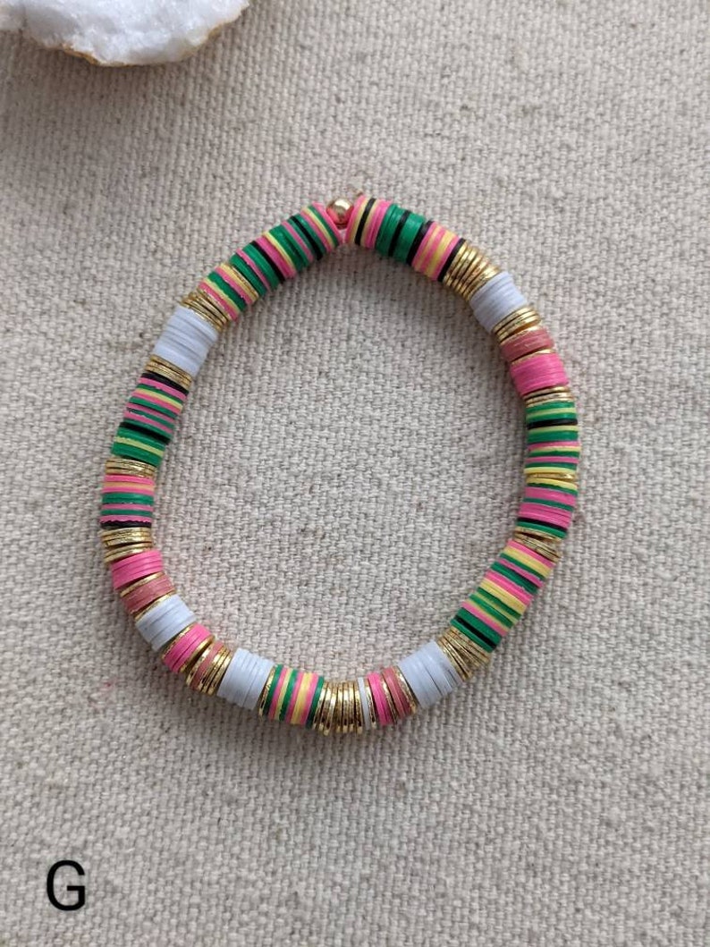 May include: A colorful beaded bracelet with pink, green, white, and yellow beads. The beads are separated by gold spacers. The bracelet is on a white fabric background.