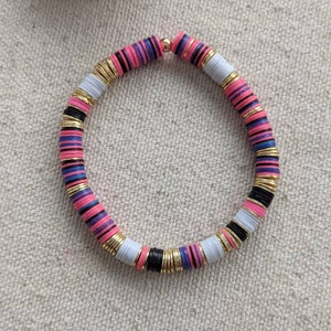 May include: A colorful bracelet made of stacked, round beads. The beads are pink, white, black, and blue with gold accents. The bracelet is on a light beige fabric background.