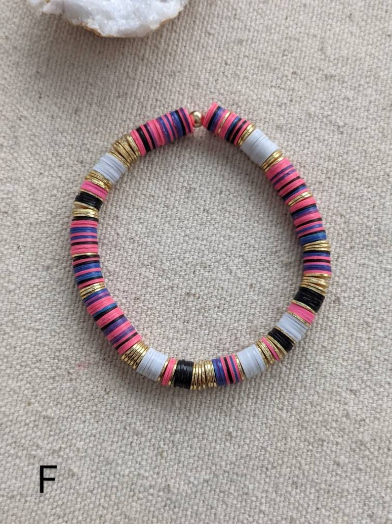 May include: A colorful bracelet made of stacked, round beads. The beads are pink, white, black, and blue with gold accents. The bracelet is on a light beige fabric background.