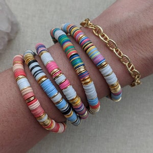 May include: A stack of colorful bracelets with white and gold accents. The bracelets are made of small, round beads and are stacked on a person's wrist.