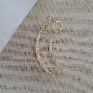 May include: Two gold crescent moon charms with a row of small clear diamonds around the edge. The charms are on a white background.