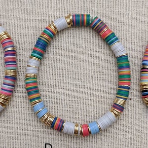 May include: A colorful beaded bracelet with white, blue, green, red, yellow, and pink beads. The beads are separated by gold spacers. The bracelet is on a beige fabric background.