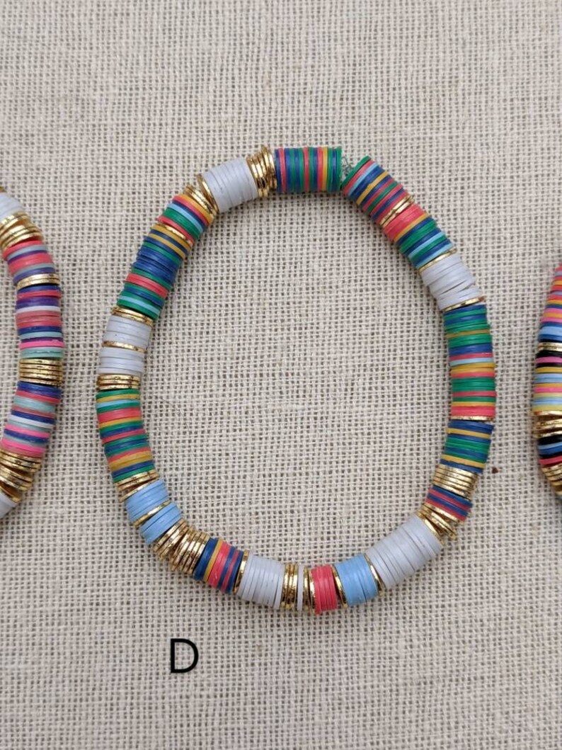 May include: A colorful beaded bracelet with white, blue, green, red, yellow, and pink beads. The beads are separated by gold spacers. The bracelet is on a beige fabric background.