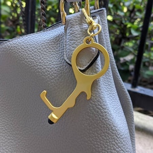 May include: A gold-colored key chain tool with a loop and a stylus tip. The tool is attached to a light gray purse.