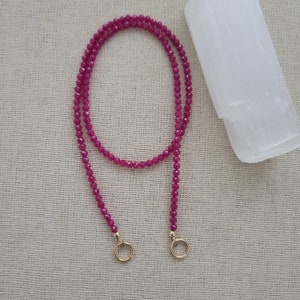 May include: A long, fuchsia-colored beaded necklace with gold-colored circular clasps. The necklace is draped on a neutral-colored surface. A white, rectangular crystal is in the background.