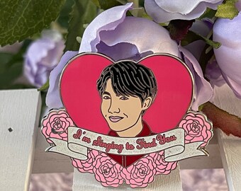Puede incluir: Un pin de esmalte en forma de corazón rosa con una ilustración de dibujos animados de una persona con cabello negro y una camisa roja. El pin tiene una cinta blanca con el texto "I'm Singing to Find You" en letras negras. El pin está rodeado de rosas rosas.