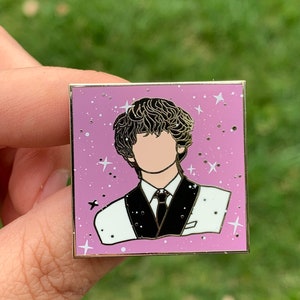May include: A gold-toned enamel pin featuring a cartoon portrait of a person with brown curly hair, wearing a white shirt and black suit jacket with a black tie. The background is a light purple with white stars.