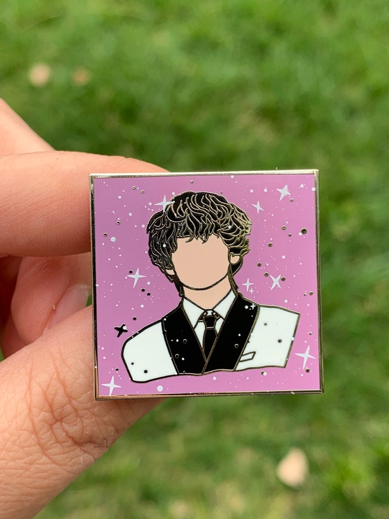 May include: A gold-toned enamel pin featuring a cartoon portrait of a person with brown curly hair, wearing a white shirt and black suit jacket with a black tie. The background is a light purple with white stars.