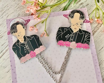 Puede incluir: Dos pines de esmalte negro con un personaje de dibujos animados con cabello negro y una chaqueta negra. Los pines están conectados por una cadena plateada con un dije de cristal rosa. El fondo es una tarjeta morada con estrellas blancas y el texto "¡Gracias por agregarme a tu colección!"