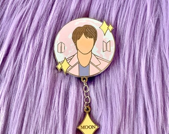 May include: A gold-colored enamel pin featuring a cartoon portrait of a person with brown hair. The pin is round and has a pink and white background with the letters "BT" on either side of the portrait. The pin has a gold chain attached to a yellow diamond-shaped charm with the word "MOON" written on it.
