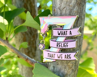 May include: A pink and white enamel pin with a rainbow swirl design. The pin features a banner with the text "What a relief that we are".