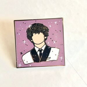 May include: A purple enamel pin with a white outline featuring a cartoon illustration of a person with dark hair wearing a black and white suit against a background of white stars.