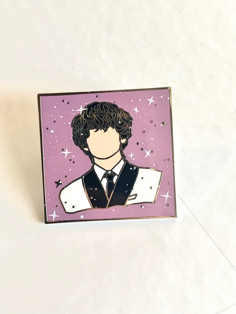 May include: A purple enamel pin with a white outline featuring a cartoon illustration of a person with dark hair wearing a black and white suit against a background of white stars.