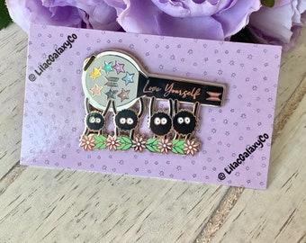 May include: A silver and black enamel pin featuring a cartoon microphone with a rainbow star design and the text "Love Yourself". Four black cartoon sheep are holding the microphone. The pin is on a purple background with white stars.