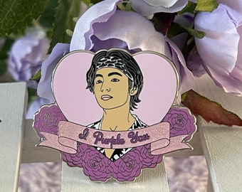 May include: A purple heart-shaped enamel pin with a cartoon illustration of a person with black hair wearing a bandana and a black and white patterned shirt. The pin has a banner with the text "I Purple You" and is surrounded by purple roses.