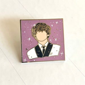 May include: A gold-colored enamel pin featuring a cartoon illustration of a person with brown hair wearing a white suit with a black tie and a white shirt. The background is a purple color with white stars.