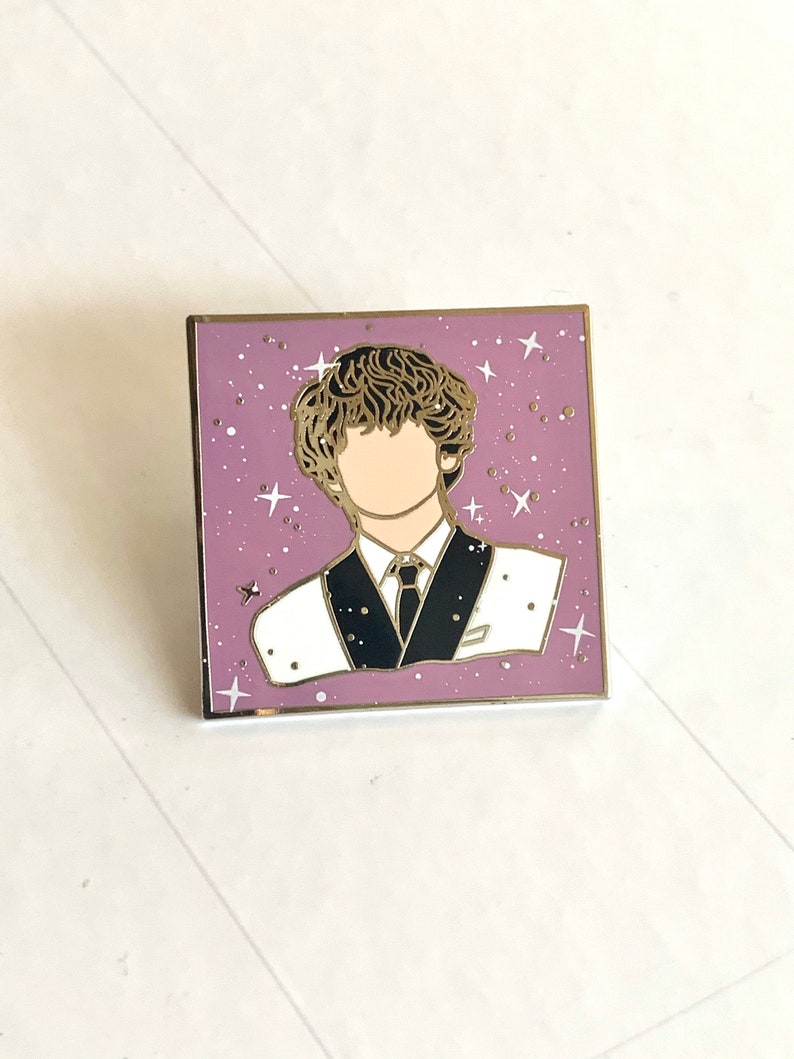 May include: A gold-colored enamel pin featuring a cartoon illustration of a person with brown hair wearing a white suit with a black tie and a white shirt. The background is a purple color with white stars.