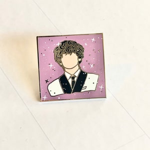 May include: A gold-toned enamel pin featuring a cartoon illustration of a person with curly hair wearing a black and white suit against a pink background with white stars.