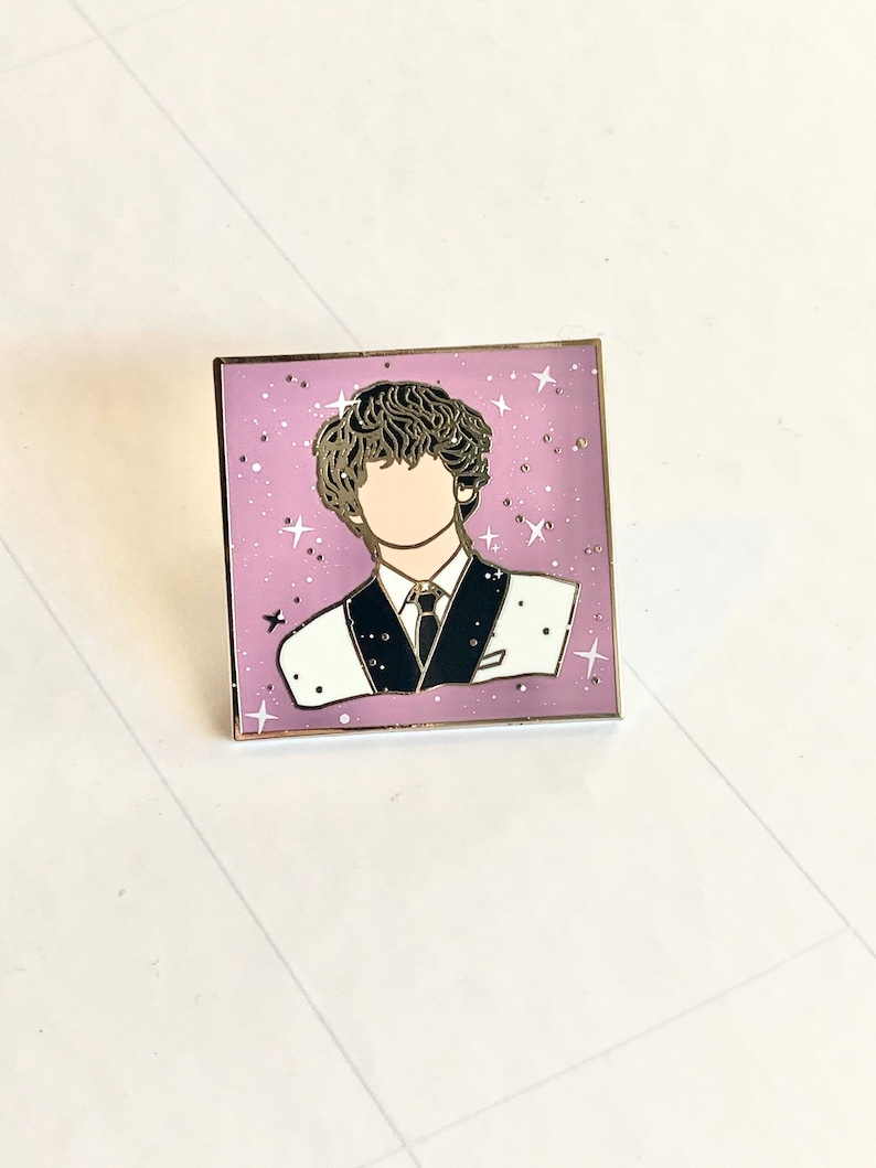 May include: A gold-toned enamel pin featuring a cartoon illustration of a person with curly hair wearing a black and white suit against a pink background with white stars.