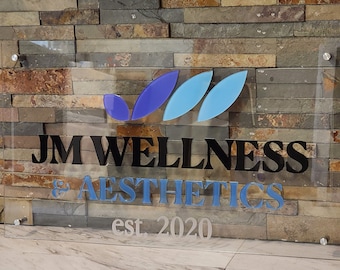 May include: A clear acrylic sign with the text "JM WELLNESS & AESTHETICS" in black and blue lettering, with a logo of three blue leaf shapes. The sign also includes the text "est. 2020" in a smaller font. The sign is mounted on a stone wall.