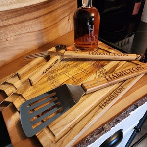 May include: A wooden cutting board with a natural edge, engraved with the name "Hamilton" and a monogram. The board is accompanied by a bottle of "Hamilton Special Reserve" whiskey, a spatula, and a set of grilling tools.