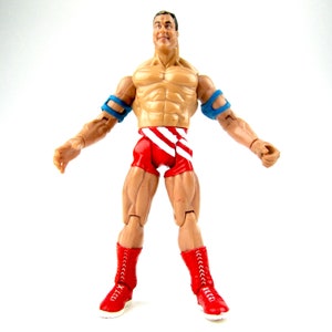 Puede incluir: Una figura de acción de lucha libre roja, blanca y azul con una construcción muscular. La figura lleva pantalones cortos rojos con rayas blancas y muñequeras azules. La figura tiene una expresión decidida en su rostro.
