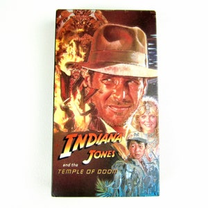 May include: A VHS tape cover for the film Indiana Jones and the Temple of Doom. The cover features a close-up of Harrison Ford as Indiana Jones, wearing his iconic fedora and a determined expression. The background features a scene from the film, with a menacing figure in the background.