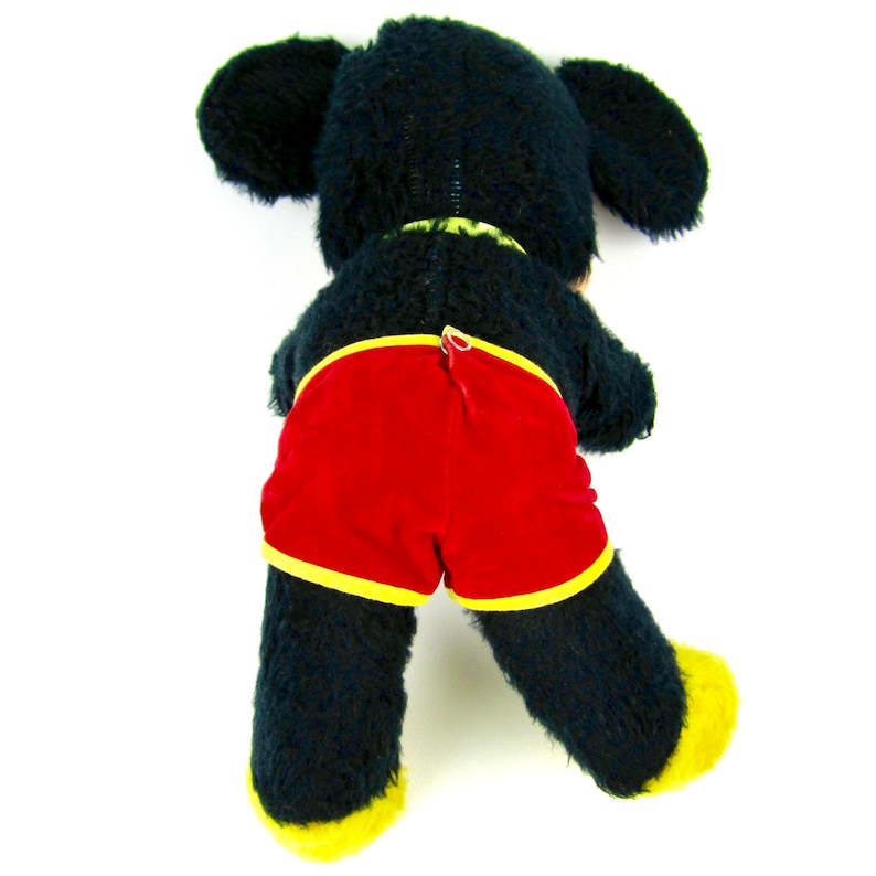 Puede incluir: Juguete de peluche negro con pies amarillos y pantalones cortos rojos. El juguete est&aacute; acostado de espaldas con la cabeza girada hacia un lado.