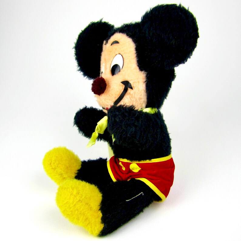 Puede incluir: Un juguete de peluche vintage de Mickey Mouse con pelaje negro, zapatos amarillos y un traje de ba&ntilde;o rojo y amarillo. El juguete tiene una expresi&oacute;n amigable con ojos negros y una nariz roja.