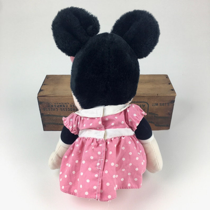 May include: A plush Minnie Mouse toy wearing a pink dress with white polka dots. The dress has a white collar and a white sash. The toy has black ears and a black face.