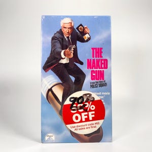 Può includere: Una videocassetta VHS del film "Una pallottola spuntata" con Leslie Nielsen. La copertina mostra Nielsen in giacca e cravatta, con una pistola, con il titolo del film in rosa. Un adesivo rosso indica uno sconto del 70%.