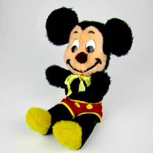 Puede incluir: Un juguete de peluche vintage de Mickey Mouse con pelaje negro, orejas grandes y zapatos amarillos. Lleva pantalones cortos rojos con lunares amarillos y una pajarita amarilla.