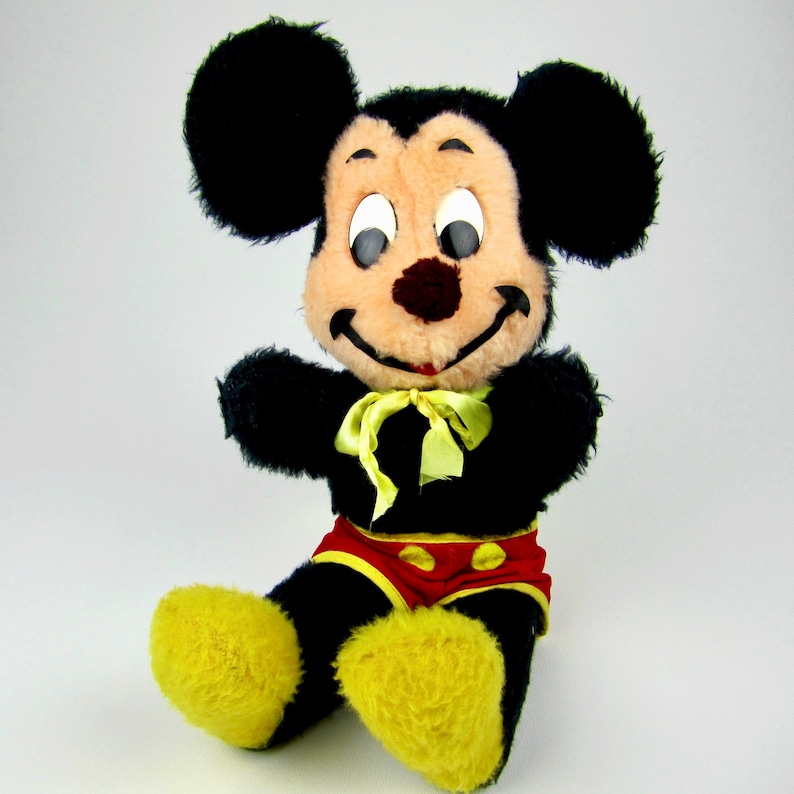 Puede incluir: Un juguete de peluche de Mickey Mouse con pelaje negro, orejas grandes y un atuendo rojo y amarillo. El juguete tiene una expresi&oacute;n amigable con ojos grandes y una amplia sonrisa.