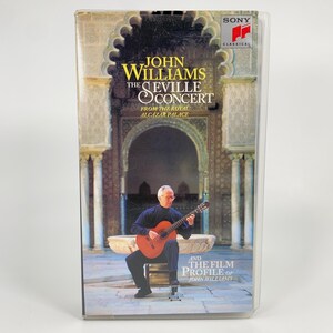 Puede incluir: Una cinta VHS de "John Williams: The Seville Concert" de Sony Classical. La portada presenta a un hombre tocando una guitarra clásica frente a una estructura de piedra. El texto en la cinta dice "John Williams The Seville Concert" y "From The Royal Alcazar Palace".
