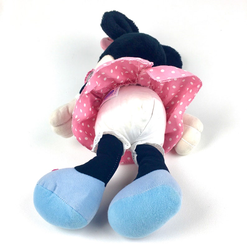 May include: A plush toy of a cartoon mouse wearing a pink dress with white polka dots and blue shoes. The toy is lying on its back with its legs in the air.