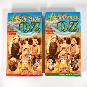 Puede incluir: Dos cintas VHS vintage con los títulos "Magic Cloak of Oz" y "His Majesty Scarecrow of Oz". Las cintas tienen portadas coloridas con imágenes del elenco de la película y el texto "L. Frank Baum's Silent Film Collection".