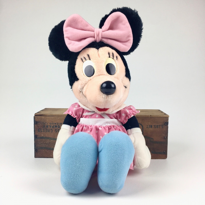 May include: A plush toy of Minnie Mouse wearing a pink dress with white polka dots and a pink bow. She has large black eyes and a black nose. She is sitting with her legs crossed and wearing blue shoes.